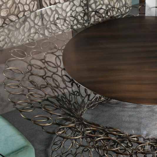 A close-up of the intricate base of the Bronze Rings Round Glass Dining Table showcases its artistic, circular design featuring interlocking bronze rings. The table rests on a textured gray rug, with part of a green chair visible in the bottom left corner.