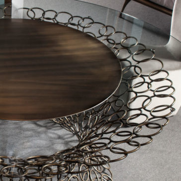 Bronze Rings Round Glass Dining Table The Bronze Rings Round Glass Dining Table features a round glass top and an intricate metal base adorned with bronze rings and circular designs. The detailed, lace-like pattern of the base radiates outward from the center. The setting is completed with upholstered chairs and a textured rug beneath the table.