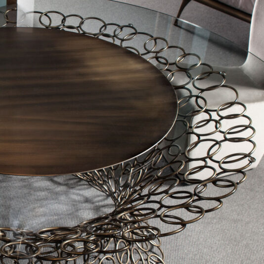The Bronze Rings Round Glass Dining Table features a round glass top and an intricate metal base adorned with bronze rings and circular designs. The detailed, lace-like pattern of the base radiates outward from the center. The setting is completed with upholstered chairs and a textured rug beneath the table.