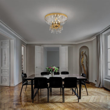 Classic Ceiling Light With Crystal Pendants A dining room features a dark wooden table surrounded by eight black chairs on a herringbone-patterned wooden floor. The Classic Ceiling Light With Crystal Pendants hangs from the ceiling, illuminating the space. A vase with flowers adorns the table, while large windows and intricate white doors brighten the room.