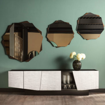 Contemporary 4 Door Sideboard A modern interior showcases a Contemporary 4 Door Sideboard in black and white set against a green wall. The sideboard features a cut-out section displaying glass items. Above, three asymmetrical mirrors reflect various parts of the room. A dark vase with white flowers sits elegantly on the sideboard.