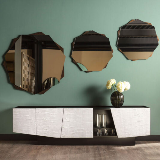 A modern interior showcases a Contemporary 4 Door Sideboard in black and white set against a green wall. The sideboard features a cut-out section displaying glass items. Above, three asymmetrical mirrors reflect various parts of the room. A dark vase with white flowers sits elegantly on the sideboard.