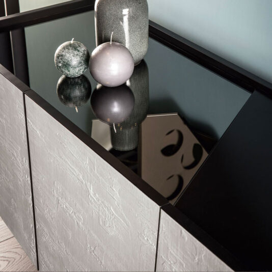 A sleek, **Contemporary 4 Door Sideboard** with a black glass top is adorned with decorative items, including a round green textured candle, a spherical gray candle, and two grey vases. The smooth, reflective surface of the glass top shows partial reflections of the objects.