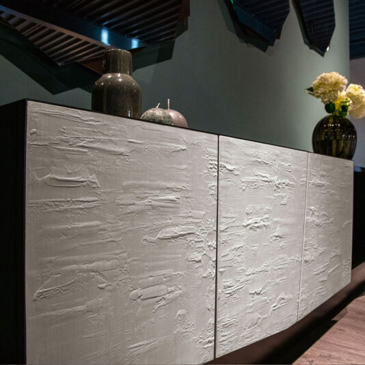 The Contemporary 4 Door Sideboard features a textured white front panel and is stylishly adorned with decorative items such as vases and candles. It stands against a dark green wall accented with angular mirrors, while one end showcases a vase filled with white flowers.