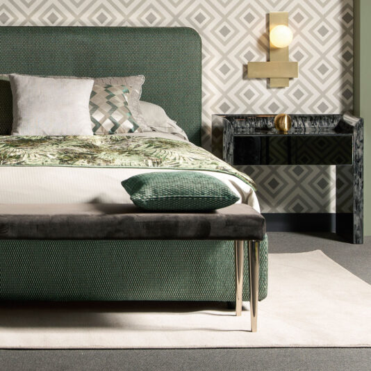 A contemporary bedroom features the Contemporary Curved Upholstered Bed, showcasing a green finish with a patterned headboard and a matching bench at the foot. The bedding is adorned with green and white leaf patterns along with geometric pillows. A black side table holds a gold decorative item, all illuminated by a stylish wall sconce.