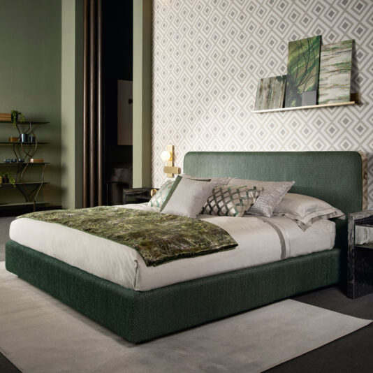 Modern bedroom showcasing a Contemporary Curved Upholstered Bed adorned with white and green bedding. The room features patterned wallpaper, a shelf with decorative items, and a small nightstand with lamp. A corner shelf with books and plants is visible in the background. Soft lighting enhances the ambiance.