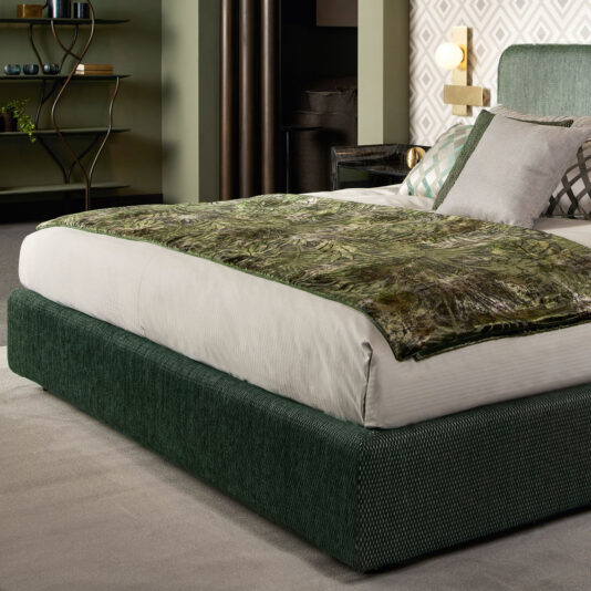 A contemporary bedroom showcasing the Contemporary Curved Upholstered Bed adorned with a white mattress and a green patterned throw blanket. The bed is accessorized with several pillows in shades of white and gray. The room features geometric patterned wallpaper, a corner shelf, and a minimalist reading lamp.