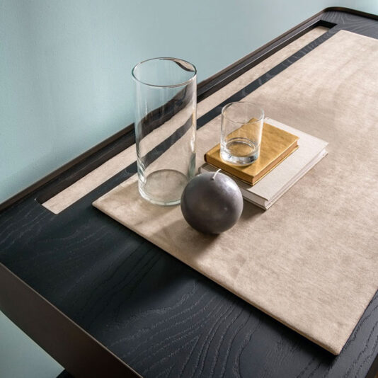 A Contemporary Designer Desk featuring a beige mat elegantly showcases a tall glass vase, a short glass cup, a stack of books topped with one that has a gold cover, and a dark spherical object. This arrangement is set against the backdrop of a light blue wall.