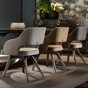 Three Contemporary Designer Dining Chairs with curved backs and cushioned seats in different neutral tones are neatly aligned beside a dark wooden table. In the background, a decorative console with two circular handles is complemented by potted plants.