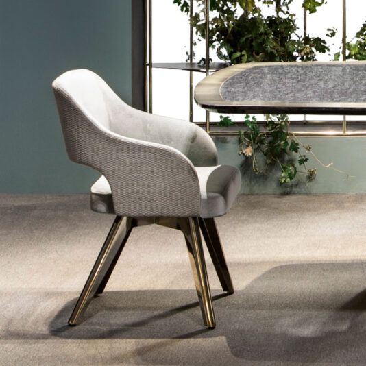 A Contemporary Designer Dining Chair with a curved back and upholstered light gray body stands on a light brown carpet. With dark wooden legs, it is positioned near a table with a gray surface, and in the background, a decorative indoor plant is visible against a window.