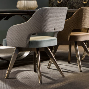 A close-up of two Contemporary Designer Dining Chairs showcasing their elegant curved backs and sleek metallic legs. The chair in the foreground is upholstered in light grey, whereas the one behind it features a beige finish. Both chairs are set on a carpeted floor beside a dark, reflective dining table.