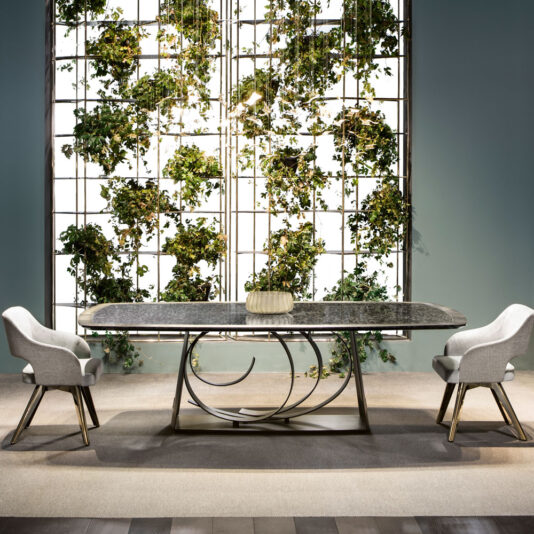 A modern dining area showcasing a long, marble-top table with an intricate metal base, flanked by two Contemporary Designer Dining Chairs. Behind the table is a large white grid filled with lush green plants, creating a striking vertical garden against a muted blue wall.