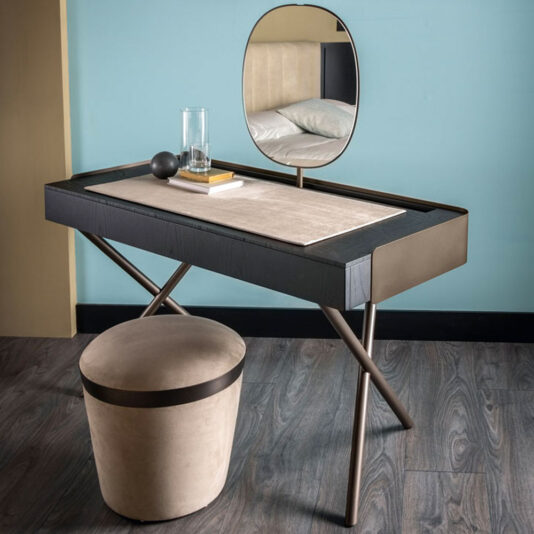 The Contemporary Dressing Table presents a modern setup with a sleek black and beige surface supported by crossed metal legs. It comes with a matching circular stool and an elegant oval mirror. The table is adorned with a small book, glass of water, and a spherical object, perfectly complementing the room's wooden floor and light blue walls.