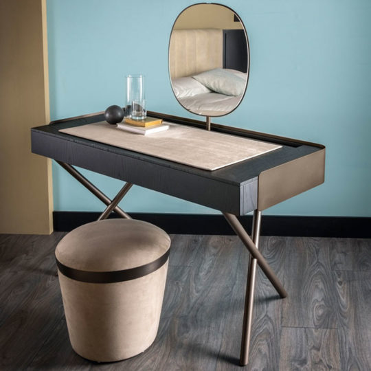 The Contemporary Dressing Table presents a modern setup with a sleek black and beige surface supported by crossed metal legs. It comes with a matching circular stool and an elegant oval mirror. The table is adorned with a small book, glass of water, and a spherical object, perfectly complementing the room's wooden floor and light blue walls.