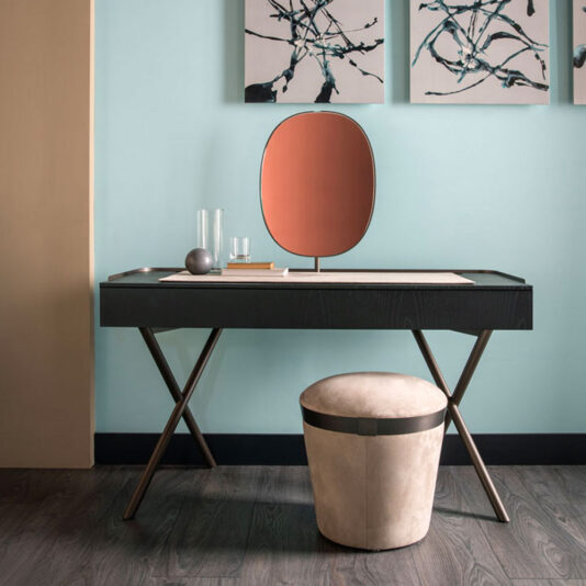 The Contemporary Dressing Table setup includes a sleek black table with crossed legs and a circular mirror with an orange hue on top. A minimalistic stool featuring a beige cushioned seat is positioned below the table. The background showcases three abstract art pieces against a light blue wall.
