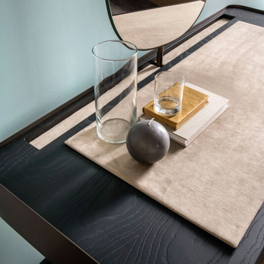 A close-up of the Contemporary Dressing Table setup featuring a tall glass vase, a small tumbler, a stack of books, and a decorative grey spherical object. These items are elegantly arranged on a beige mat atop the dark wooden surface with a partial view of the mirror in the background.