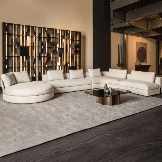 A modern living room showcasing the Contemporary Leather Modular Corner Sofa with a round chaise lounge, elegantly set on a light gray carpet. Behind the sofa stands a bookshelf designed with geometric patterns and integrated lighting, while minimalist decor accents the space and dark flooring completes the sophisticated look creating a sociable living room
