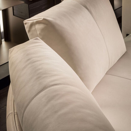 Close-up of the Contemporary Leather Modular Corner Sofa in beige leather, featuring a plush cushion and headrest. The smooth and comfortable upholstery showcases subtle stitching details. The background reveals parts of wooden furniture and ambient indoor lighting.
