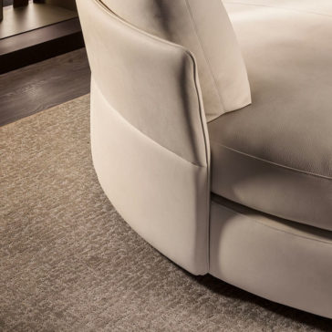 A close-up view of a beige, circular Contemporary Leather Modular Corner Sofa. The sofa is positioned on a light brown carpet, and the image focuses on the side of the sofa, highlighting its smooth upholstery and seamless stitching.