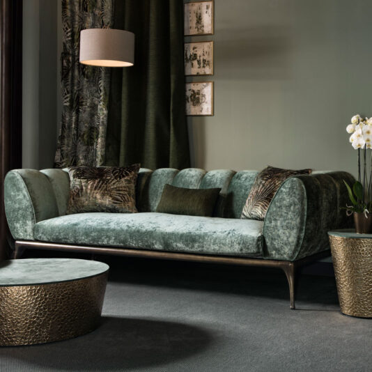 Contemporary Luxury Sofa A luxurious living room featuring a plush, green velvet Contemporary Luxury Sofa adorned with decorative pillows. Above the sofa, three framed art pieces are displayed alongside a hanging lamp. The space includes a round, textured coffee table and a potted orchid plant, all set against muted green walls.