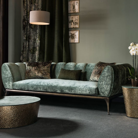 A luxurious living room featuring a plush, green velvet Contemporary Luxury Sofa adorned with decorative pillows. Above the sofa, three framed art pieces are displayed alongside a hanging lamp. The space includes a round, textured coffee table and a potted orchid plant, all set against muted green walls.