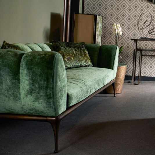 Contemporary Luxury Sofa A Contemporary Luxury Sofa in green velvet, adorned with rounded armrests and wooden legs, is situated in the living room. The sofa includes two green patterned cushions. Surrounding it are geometric patterned wallpaper, a tall mirror, a small table with a white orchid, and an elegant floor accent piece.