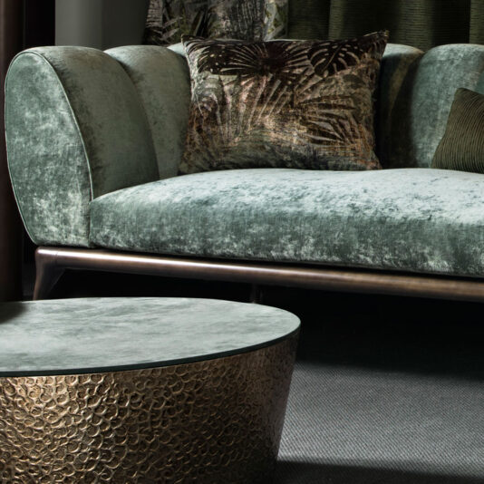 Contemporary Luxury Sofa A stylish living room setting featuring the Contemporary Luxury Sofa in a luxurious green velvet, paired with a metallic patterned throw pillow. In the foreground, there's a round coffee table with a textured, gold-toned base and a matching green velvet top. The contemporary ambiance is modern and elegant.
