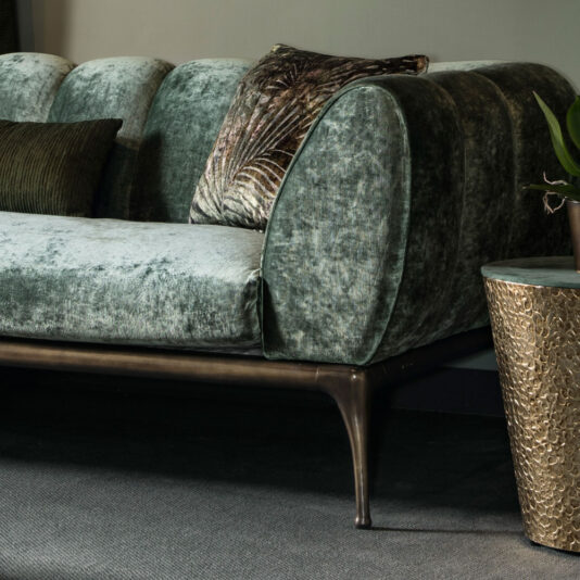 Contemporary Luxury Sofa The Contemporary Luxury Sofa, upholstered in luxurious green velvet and featuring tufted backrests and sleek metal legs, is elegantly placed on a dark carpet. The sofa is complemented by a patterned throw pillow and a ribbed cushion for added comfort. Beside it stands an elegant metallic side table adorned with a potted plant.
