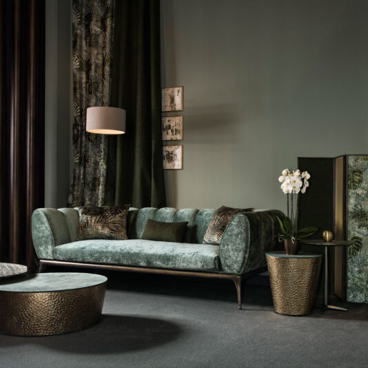 Contemporary Luxury Sofa A stylish living room showcases the Contemporary Luxury Sofa, upholstered in plush green velvet and adorned with patterned cushions. A round, textured coffee table and a side table featuring a white orchid enhance the decor. Dark green curtains, wall art, and a hanging lamp complete the room's sophisticated look.