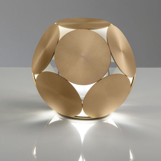 A Contemporary Metal Disc LED Table Lamp is elegantly positioned on a white surface, exhibiting an intricate geometric design featuring large, overlapping circular metal plates with a brass finish. Soft light streams through the gaps between the plates, enhancing its stylish and modern aesthetic.