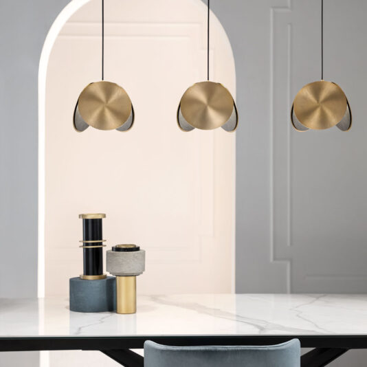 A modern interior scene features three Contemporary Metal Disc Pendant Lights with gold circular designs above a white marble dining table. The table is adorned with artistic cylindrical objects in black, gold, and grey shades, and there's a grey chair in front of the table.