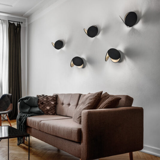 Contemporary Metal Disc Wall Light A modern living room featuring a brown sofa with pillows and a blanket, positioned against a white wall adorned with five Contemporary Metal Disc Wall Lights. Natural light streams in from a window with dark curtains. A black coffee table sits in front of the sofa.