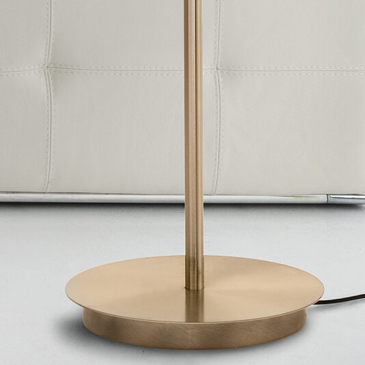 A close-up of the base of the Contemporary Metal Disc LED Floor Lamp, showcasing its sleek design and brushed gold finish, as it stands on a white surface. The background features white tufted upholstery, possibly part of a chair or sofa. A black power cord is partially visible.