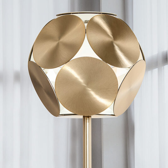 The Contemporary Metal Disc LED Floor Lamp features a spherical shade made of overlapping golden metal discs, offering a sleek geometric design. Positioned in front of a light-colored, sheer curtain, this modern LED floor lamp enhances any space with its sophisticated style.