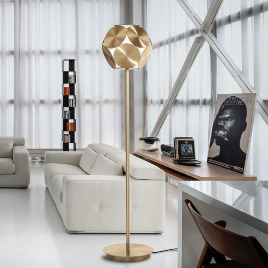 A modern living room with a tall, gold-colored Contemporary Metal Disc LED Floor Lamp featuring a geometric design. The space includes a white sofa, a small shelving unit filled with books, a wooden desk adorned with decorative items, and a wall poster. Natural light floods the room through large floor-to-ceiling windows.