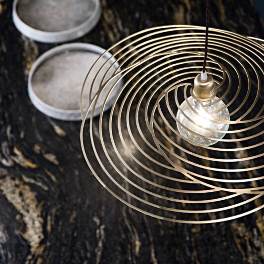 Contemporary Spiral Chandelier A close-up of the Contemporary Spiral Chandelier showcases its concentric circular metal rings surrounding a glowing bulb in the center. The background displays a dark, textured surface with an intriguing pattern, accompanied by two round, shallow bowls topped with stone-like lids.