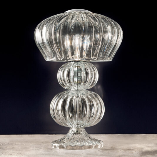 The Contemporary Tiered Glass Table Lamp features an elegant, dome-shaped glass shade and a base made up of three stacked, spherical glass elements. Its intricately ridged design produces a glowing, shimmering effect when placed against a dark background.