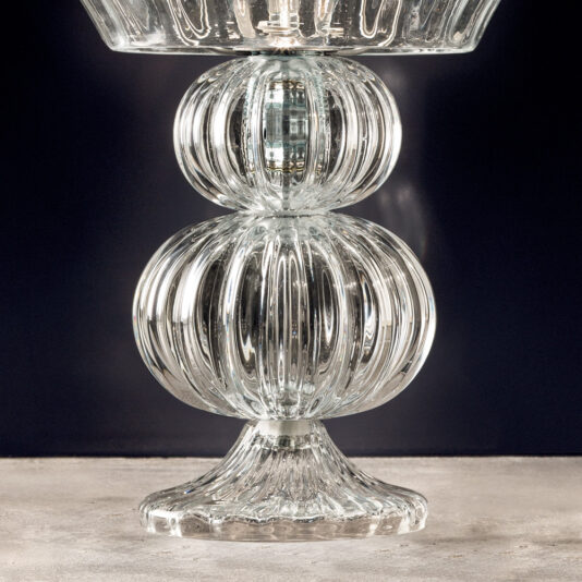 A close-up view of the Contemporary Tiered Glass Table Lamp featuring a ribbed design. The lamp's structure includes a wide conical base, a central spherical segment, and an upper flared bowl-like section, all crafted from clear glass. The dark background accentuates the elegant glass design.