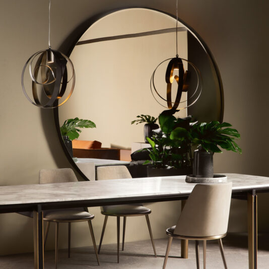A modern dining room featuring the Contemporary Twisted Bronze Round Mirror above a rectangular dining table with a marble top. Three beige upholstered chairs are placed around the table. Two spherical pendant lights hang above, and a potted plant adds natural charm to the scene.