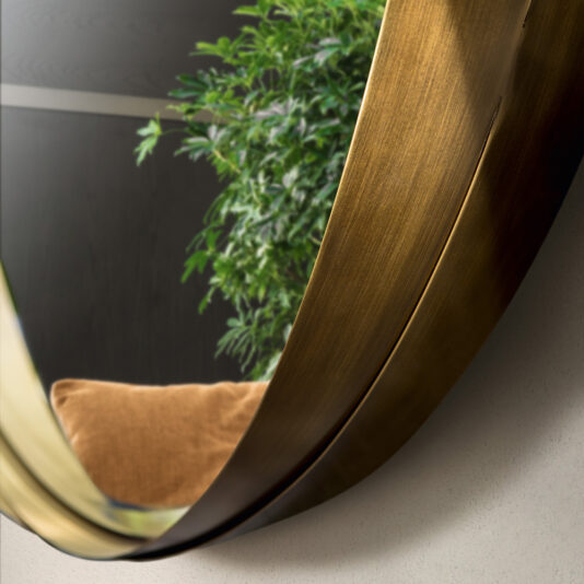 A close-up image of the Contemporary Twisted Bronze Round Mirror. The mirror reflects a portion of a green leafy plant and a brown cushion or chair against a dark backdrop, creating a modern and cozy ambiance.