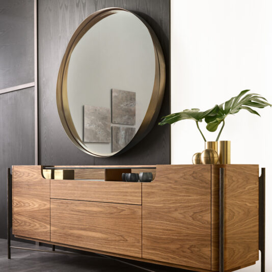 A modern interior showcases a **Contemporary Twisted Bronze Round Mirror** mounted on a dark wall above a sleek wooden sideboard. The sideboard is adorned with minimalistic decor items, including golden vases filled with green leaves, while artwork graces the background.
