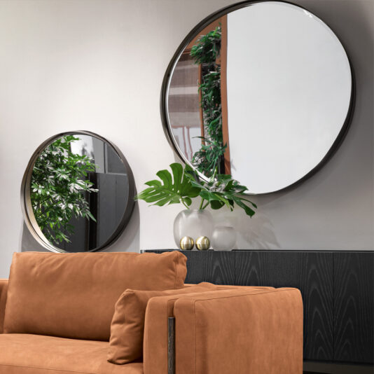 A modern living room featuring a brown leather couch in the foreground. Behind the couch, a black console table is adorned with two Contemporary Twisted Bronze Round Mirrors. One mirror reflects greenery, and on the table sits a vase with a large green leaf and two small spheres.
