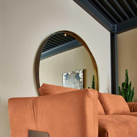 A Contemporary Twisted Bronze Round Mirror with a thin, dark frame is mounted on a beige wall. Below it is a modern, orange suede sofa. There is a small square abstract painting in muted colors on the wall next to a tall, green cactus plant. A dark metal ceiling structure is partially visible.