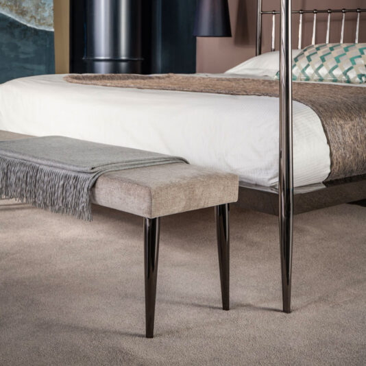 Contemporary Upholstered Bench An elegant bedroom showcases a modern bed complete with a white mattress, patterned pillows, and a brown textured throw. The focal point at the foot of the bed is the Contemporary Upholstered Bench, featuring a plush beige cushion and dark metallic legs, beautifully adorned with a neatly folded gray fringed blanket.