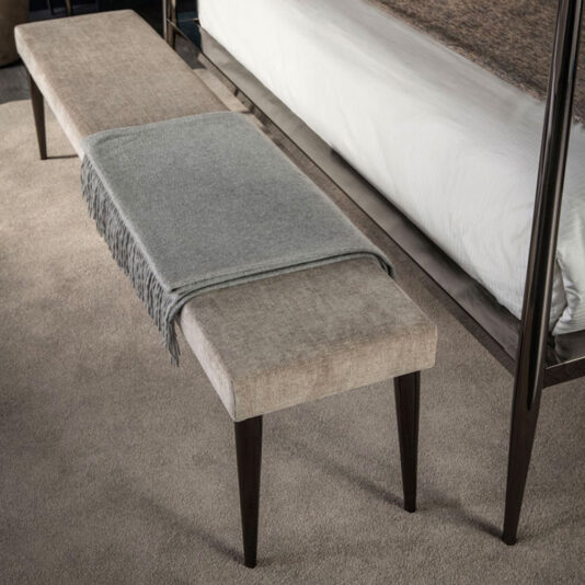 The Contemporary Upholstered Bench with dark wooden legs is positioned at the foot of a bed adorned with a white comforter. A light gray throw blanket is elegantly draped over the center of the bench, enhancing the cozy atmosphere of the carpeted room.