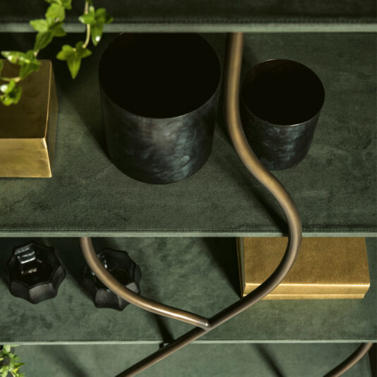 Contemporary Wrought Iron Bookcase A close-up of a decorative shelf on a green surface, featuring dark cylindrical containers, a twisted brass rod, and two gold rectangular boxes. Small green plants frame the scene, adding a touch of nature to the elegant display on the Contemporary Wrought Iron Bookcase.