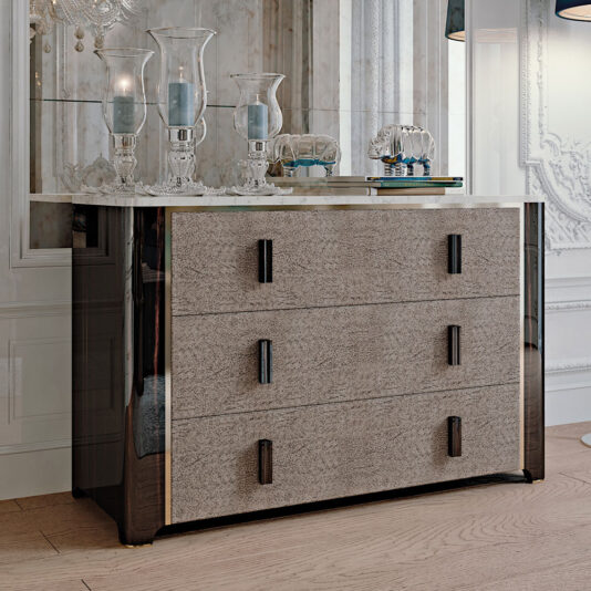 The Elegant Ebony Chest of Drawers, with its modern and stylish design, features a reflective dark glossy surface complemented by three textured beige drawers. Decorative glass candle holders and small ornamental items adorn it. This exquisite piece is set in a luxurious room with light-colored walls and intricate detailing.