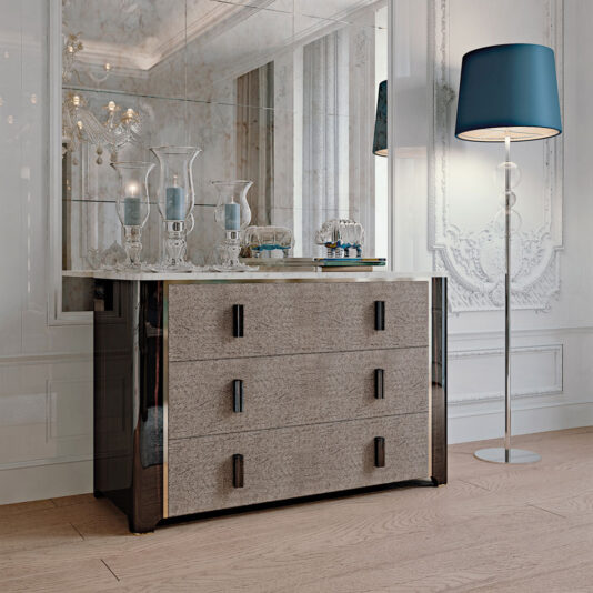 A modern, elegant living room features the Elegant Ebony Chest of Drawers with a reflective surface and three drawers, adorned with vases and trays. A tall floor lamp with a blue lampshade stands nearby. The space has ornate wall moldings, a large mirror, and wooden flooring.