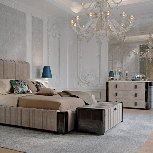 A luxurious bedroom featuring an upscale bed with a beige upholstered frame, topped with decorative pillows and a beige blanket. A grand crystal chandelier hangs from the ceiling, and an ornate mirror sits above the Elegant Ebony Chest of Drawers. The room boasts elegant wall paneling and a plush area rug.