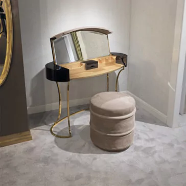 Classic Ebony Dressing Table Positioned gracefully on a beige carpeted floor, the Classic Ebony Dressing Table, featuring a folding mirror, black and wood accents, gold curved legs, and multiple compartments, is complemented by a round plush ottoman upholstered in matching beige fabric.
